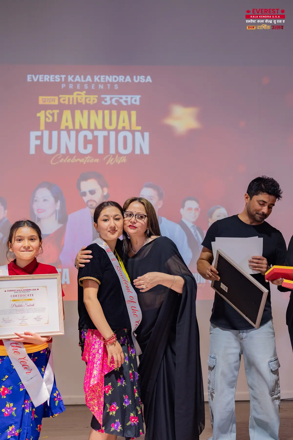 A young EKK student holds her certificate alongside two adults on stage