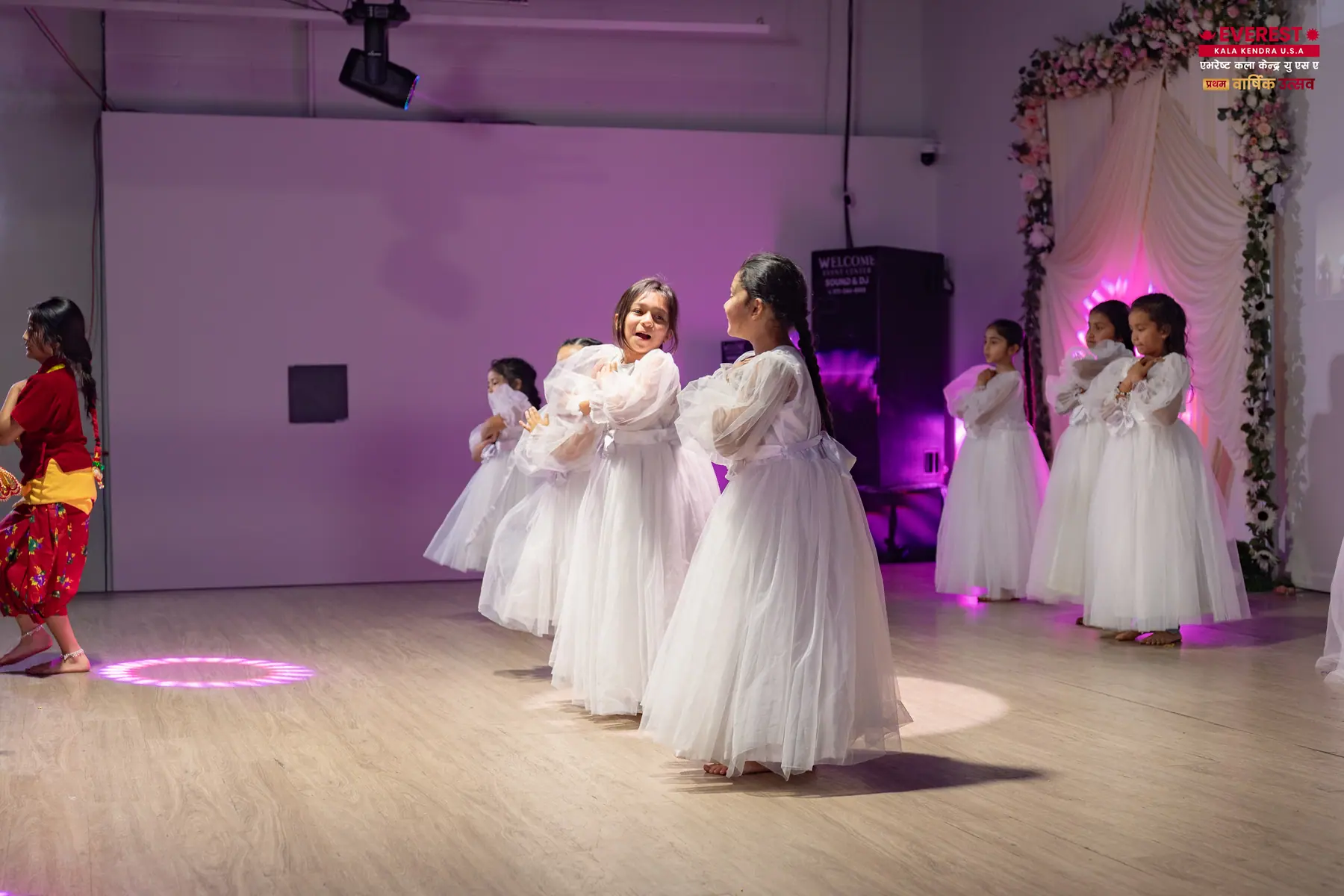Young EKK dancers performing in white dresses lit by purple and pink stage lighting