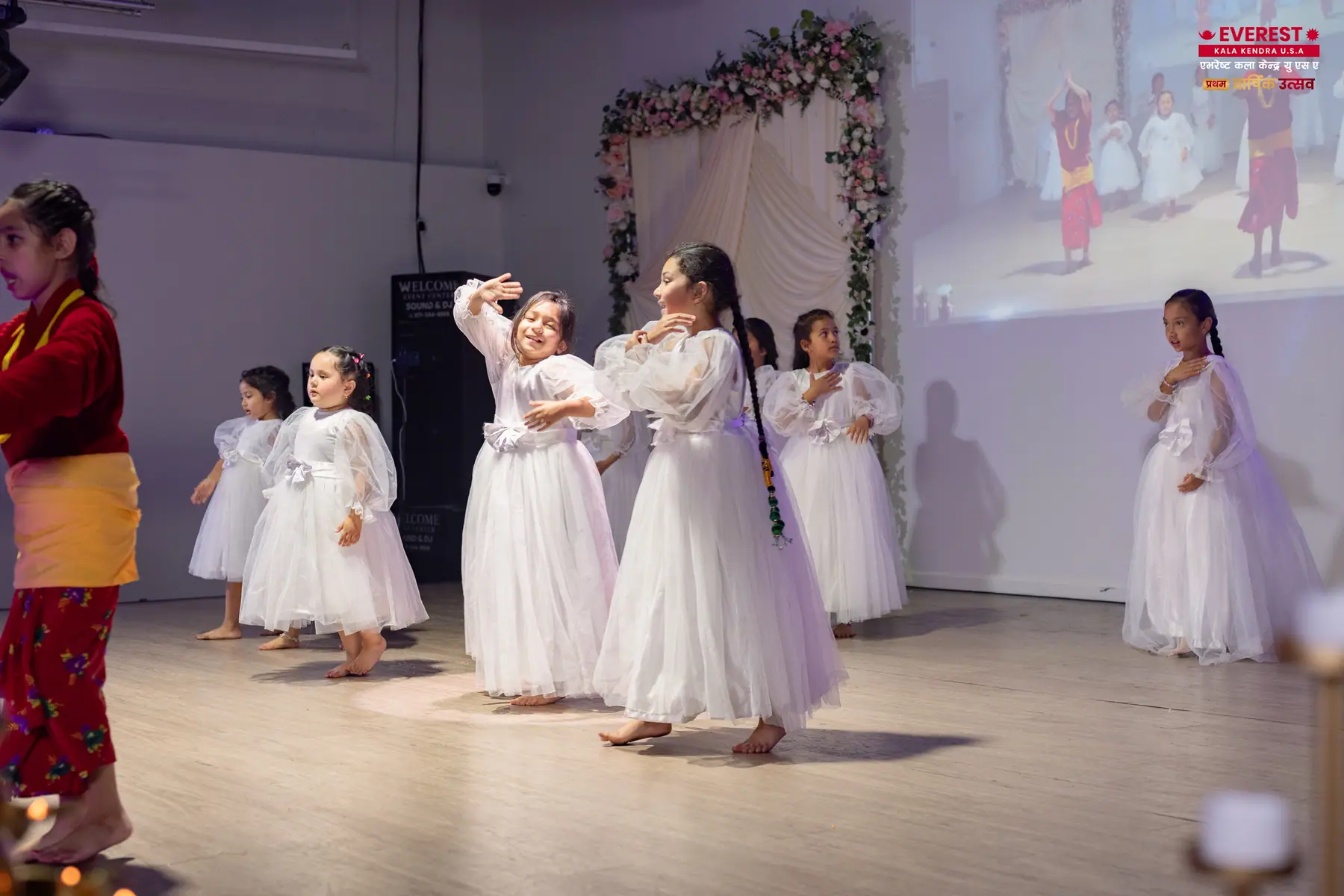 Young EKK dancers on stage beside an adult in traditional Nepali dress guiding the routine