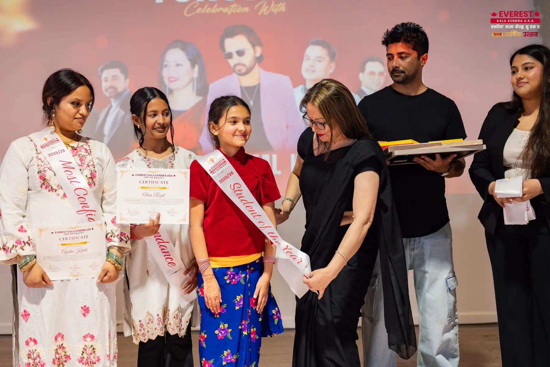 An EKK student wearing a ceremonial sash receives a certificate on stage