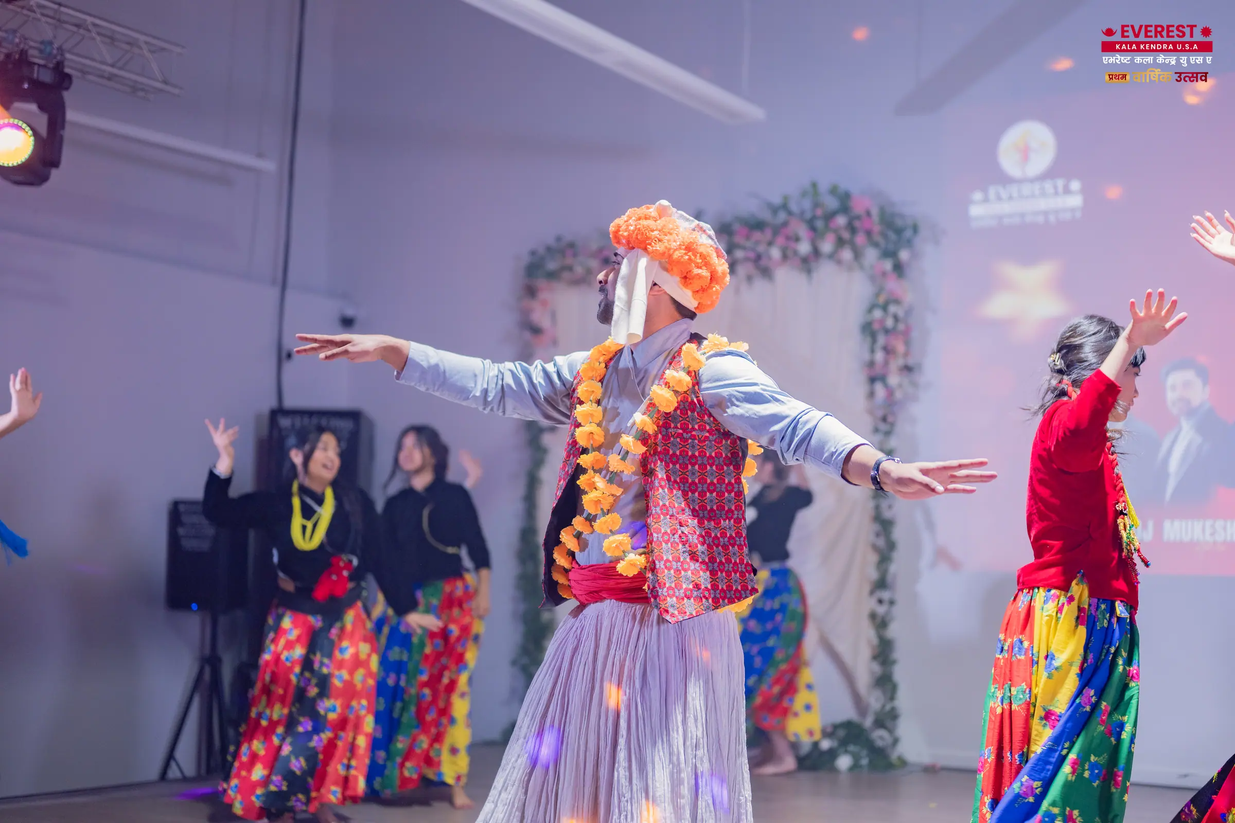 An EKK dancer in a brightly striped traditional Nepali costume and feathered headdress performs on stage with arms outstretched